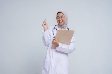 Beyaz önlüklü ve gözlüklü Asyalı kadın doktor. Elinde steteskop, elinde pano, kameraya bakıyor, gülümsüyor ve bir şeyler düşünüyor.