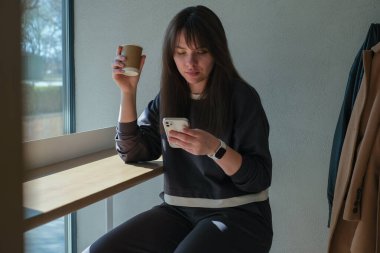 Bir kadın bir elinde kahve fincanıyla tabureye oturmuş akıllı telefonunu kontrol ediyor. Güneş ışığı pencereden akar, rahat atmosferini aydınlatır ve rahat çevresini aydınlatır..