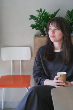 Bir kadın bir kafede, elinde bir fincan kağıt kahveyle rahatça oturuyor. İçerideki bitkiler ve modern mobilyalarla çevrili olan mesafeye dikkatle bakıyor..