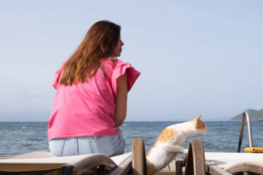 Bir kişi şezlongda oturur ve denizin huzurlu manzarasının tadını çıkarır. Neşeli bir kedi yakınlarda geziniyor, plaj atmosferine büyüleyici bir dokunuş ekliyor..