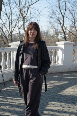 Genç bir kadın şehir parkında güvenli bir şekilde dolaşır, modern bir kıyafet giyer ve çıplak ağaçlarla çevrilidir. Açık gökyüzü sahnede sakin bir atmosfer yaratıyor..