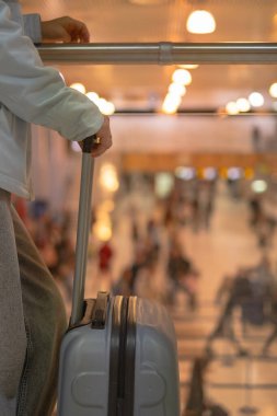 Gezgin, elinde bavulla bir tırabzanın yanında duruyor. Yoğun havaalanı terminalini izliyor..