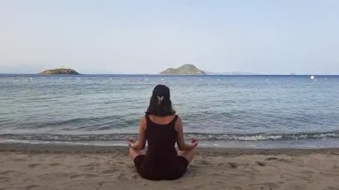 Kadın, gün doğumunda sakin bir plajda derin dalgalar ve uzak adalarla meditasyon yapıyor.