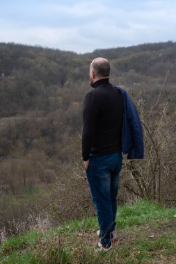 Bir adam ağaçlar ve yapraklarla kaplı uzak tepelere dikkatlice bakar bulutlu bir bahar gününde çimenli bir yamaçta dururken doğanın huzurunun tadını çıkarır..