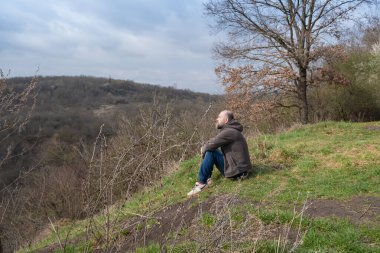 Bir kişi çimenli bir yamaçta sessizce oturur, yemyeşil yamaçlara ve bulutlu gökyüzünün altındaki ağaçlara bakar. Manzara tomurcuklanan yapraklarla ilkbaharın başlarını yansıtıyor..