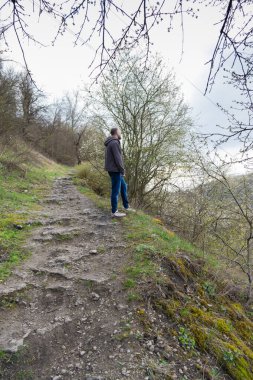 Günlük giysiler giymiş bir adam, yeşilliklerle çevrili engebeli bir yolda düşünceli bir şekilde duruyor. Gökyüzü bulutlu, yaklaşan hava değişimine işaret ediyor..