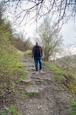 Bir adam dağılmış kayalar ve yeşilliklerle kaplı engebeli bir yolda yol alıyor. Etrafındaki ağaçlar, bulutlu gökyüzünün tepende belirmesiyle huzurlu bir ortam yaratıyor ve sakin bir açık hava deneyimi yaratıyor..