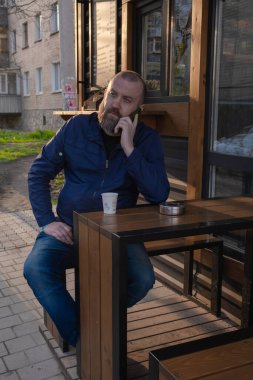 Sakallı bir adam kafenin dışındaki ahşap bir masada oturuyor ve bir telefon görüşmesi yapıyor. Işıklar öğleden sonra olduğunu gösteriyor. Arka planda birkaç bina var..