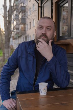 Sakallı bir adam şehir merkezinde açık bir masada oturuyor, düşünceli bir şekilde beyaz bir fincandan kahve yudumluyor. Güneş batıyor, çevreye sıcak bir parıltı saçıyor..