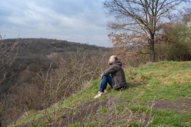 Bir adam tepenin yamacında sessizce oturur, aşağıdaki vadiyi seyreder. Manzara bereketli ama çıplak. Bu da kıştan bahara geçişi gösteriyor..