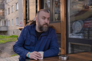 Sakallı bir adam sakince bir kafede oturuyor, elinde bir fincan..