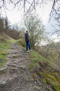 Bir adam sakin bir ifadeyle dışarıda duruyor, kahverengi ceket ve mavi kot pantolon giyiyor, sakin bir manzarayla çevrili.