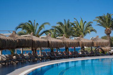 Parlak mavi bir havuzun kenarında uzanan güneşli yerler gür palmiye ağaçları ve sazdan şemsiyelerle çevrili, berrak bir gökyüzü altında rahatlamaya davetiye çıkarıyor..