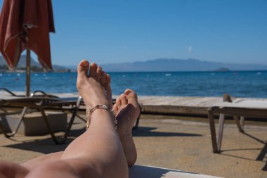 Güneşli ayaklar plajın yanındaki şezlongda dinleniyor, sakin okyanusa bakıyor. Açık mavi gökyüzü ve uzak dağlar sakin kıyı atmosferine eklenir..