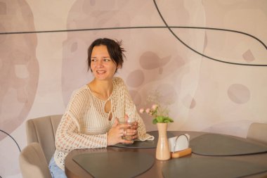 Bir kadın bir kafede şık bir masada oturuyor, elinde bir bardak içecekle. Doğal ışık boşluğu dolduruyor, modern dekoru ve onun neşeli ifadesini vurguluyor..