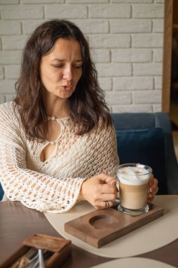 Modern bir kafede oturan bir kadın elinde buzlu bir bardak latte tutuyor. Örgü bir bluz giyiyor ve içkiyle meşgul görünüyor, sakin bir öğleden sonra atmosferinin tadını çıkarıyor..