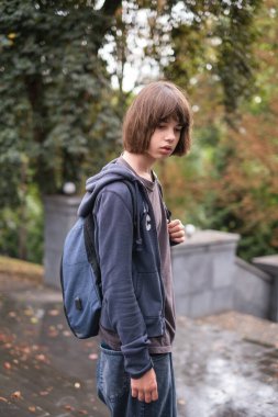 Sırt çantalı 13 yaşında bir çocuk ıslak basamaklarda durup dikkatle bakıyor. Sahne yemyeşil ve atmosfer sakin, yağmurlu bir günü yansıtıyor..