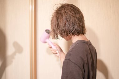 13 yaşında bir çocuk, parlak bir banyoda ıslak saçlarını şekillendirmek için saç kurutma makinesi kullanıyor. Günlük kıyafetleri ve görünüşü öğleden sonra rahatlayacağını gösteriyor..