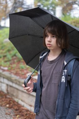 13 yaşında bir çocuk, elinde siyah bir şemsiyeyle parkta sessizce duruyor. Yağmur hafifçe yağar, yerde su birikintileri oluşturur, renkli sonbahar yaprakları ise yolu kaplar..