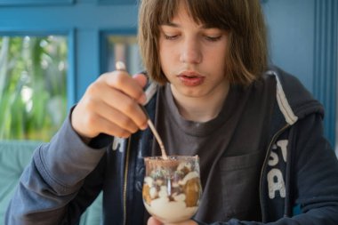 13 yaşında bir çocuk bir kafede masada oturuyor, dikkatle bardaktaki tatlıyı karıştırıyor. Sıcak atmosferde mavi duvarlar ve yeşillikler var. Keyif anı güzelleştiriyor..