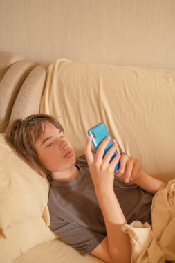 13 yaşında bir çocuk kanepede rahat bir şekilde uzanıyor, akıllı telefonunu dikkatlice kullanıyor. Sıcak, yumuşak ışık, rahat bir atmosfer yaratıyor..