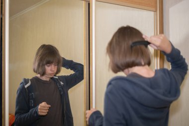 Genç bir insan aynanın önünde duruyor, saçını düşünceli bir şekilde tarıyor. Oda hafifçe aydınlatılıyor, önlerindeki güne hazırlanırken rahat bir atmosfer yaratıyor..