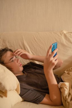 Ergen çocuk bir yatakta oturmuş, öğleden sonra rahat bir evde akıllı telefona bakıyor..