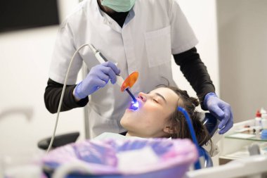 Bir diş hekimi modern bir diş muayenehanesinde hastanın dişlerini temizlemeye odaklanır. İşlem yüksek teknolojili ekipmanlarla yapılırken hasta rahat bir şekilde oturuyor..