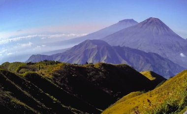 Canlı bir dağ manzarası sergiliyor yemyeşil yuvarlanan tepeler, yükselen tepeler, ve geniş, açık mavi bir gökyüzü, doğayla bir huzur ve bağlantı hissi uyandırıyor.