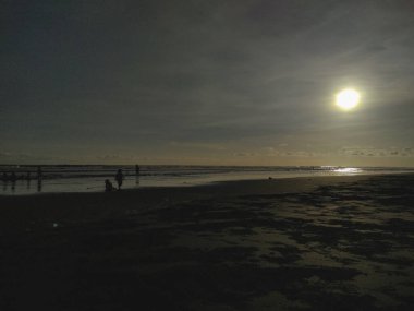 Gün batımında zengin altın renklerle sahilde yürümekten hoşlanan insanların siluetleri. Barışçıl atmosfer batan güneşin ışıltısı altında yansıyan suyun cazibesini arttırır.