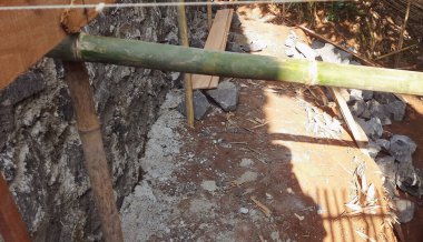 Yapısal destek veya hazırlık için kullanılan bir inşaat sahasında yerleştirilmiş bambu çubukları ve çakılların yer aldığı görüntü. Arka planda korunmasız taş duvar ve parlak güneş ışığı ayarı vurguluyor