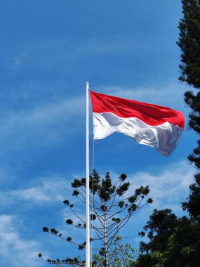 Endonezya bayrağı rüzgar tarafından dalgalanan bir direkte dalgalanıyor mavi gökyüzü ve bulutların arka planına karşı.