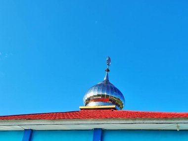 Gümüş kubbeli bir cami ve açık mavi bir gökyüzünün altında duran kırmızı bir çatı..