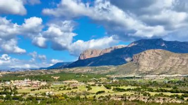 Görkemli dağların ve yemyeşil tarlaların çarpıcı manzarası kabarık bulutlarla dolu açık mavi bir gökyüzünün altında uzanır.