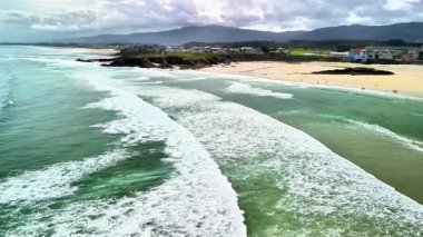 Hava görüntüsü muhteşem okyanus dalgalarının yavaşça kumlu bir sahile ulaştığını ve arka planda görkemli dağlar olduğunu gösteriyor.