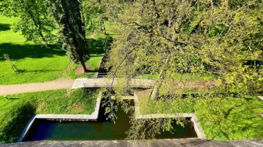Bu hava görüntüleri, yemyeşil yemyeşil ile çevrili sakin bir nehrin üzerindeki taş bir köprüyü gösteriyor. Güneş ışığı su üzerinde gökkuşağı etkisi yaratır.