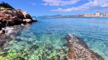 Canlı, berrak turkuaz bir su, engebeli kayalık bir sahil manzarası ve kentsel bir zemin.