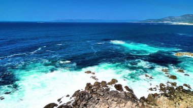 Canlı turkuaz suların çarpıcı görüntüsü sahil şeridi boyunca engebeli kayalara çarpıyor.