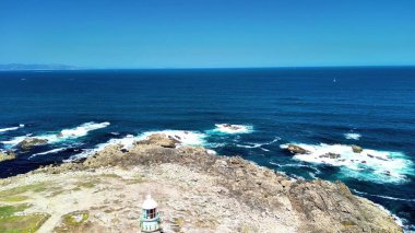 Açık mavi gökyüzü ve çarpan dalgaların altında okyanus kıyısındaki kayalık bir kıyı bölgesinin nefes kesici manzarasının tadını çıkarın.