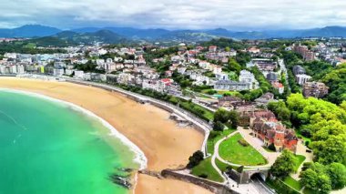 Hava manzarası, sakin bir sahil kasabası, yeşil tepeler ve modern binalar sergiliyor.