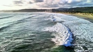 Nefes kesici kıyı manzarası, günbatımı saatinde sakin renklerle eşlik eden çarpan dalgaları içerir.