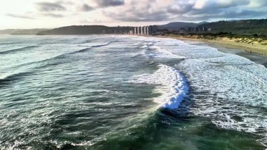 Nefes kesici kıyı manzarası, günbatımı saatinde sakin renklerle eşlik eden çarpan dalgaları içerir.