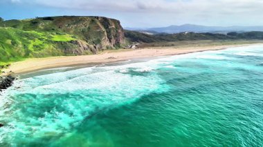 Sakin bir plajı, yumuşak dalgaları ve berrak gökyüzünün altındaki verimli uçurumları gösteren nefes kesici hava manzarası..