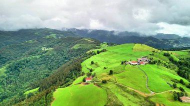 Sersemletici yeşil tepeler ve dağlarla çevrili çiftlik evleri bulutlu bir gökyüzünün altında..