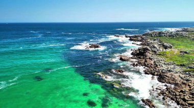 Nefes kesen hava görüntüleri, berrak mavi gökyüzünün altında kayalık kıyılarda çarpıcı bir turkuaz okyanus buluşmasını canlı bir şekilde gösteriyor.