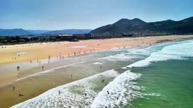 Canlı ve canlı bir plaj sahnesi. İnsanların muhteşem bir dağ zeminine karşı dalgaların tadını çıkardığı bir sahne.