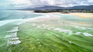 Nefes kesici bir hava manzarası bulutlu bir gökyüzünün altındaki kumlu kumsalda usulca süzülen yumuşak dalgaları yakalar.