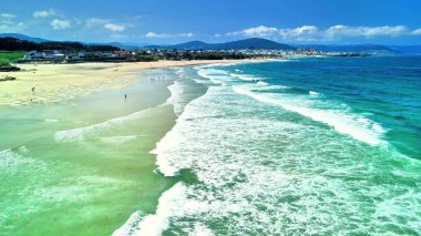 Hava manzarası, güneşli bir günde kristal berrak turkuvaz suları ve hafif dalgalarla sakin bir plajı gözler önüne seriyor.