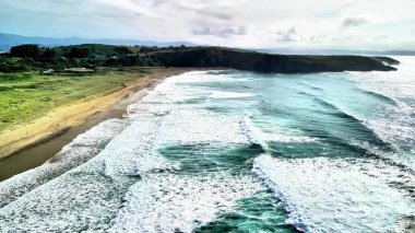 Göz kamaştırıcı kıyı manzaraları güzel bir şekilde kumlu kıyılara vuran dalgalar, çevredeki canlı yeşilliklerle güçleniyor.