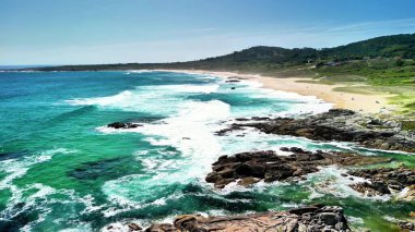 Güçlü dalgaların berrak mavi bir gökyüzünün altında sakin bir sahile çarpmasının nefes kesici ve çarpıcı görüntüsünü deneyimleyin.